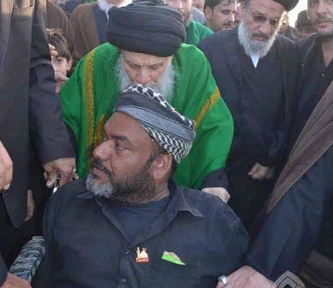 بالصورة :احد مراجع النجف يدفع عربة احد الزوار المعاقين اثناء مسيره على طريق بين النجف وكربلاء